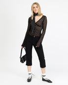 Model trägt eine schwarzes Langarmshirt aus zarter Spitze mit V-Ausschnitt und Brustabnähern zu einer schwarzen Caprihose und schwarzen Schnür Ballerinas und einer schwarzen Tasche sowie ein Polkadot Tuch.