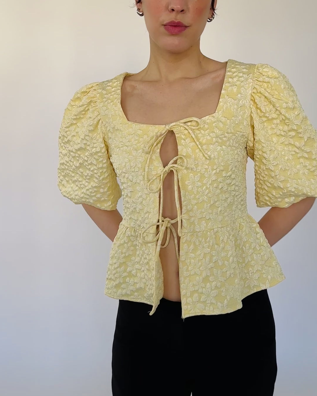 Model trägt eine voluminöse, buttergelbe Kurzarmbluse mit floralen Details, Bindebändern und Peplum-Schnitt zu einer schwarzen Caprihose.