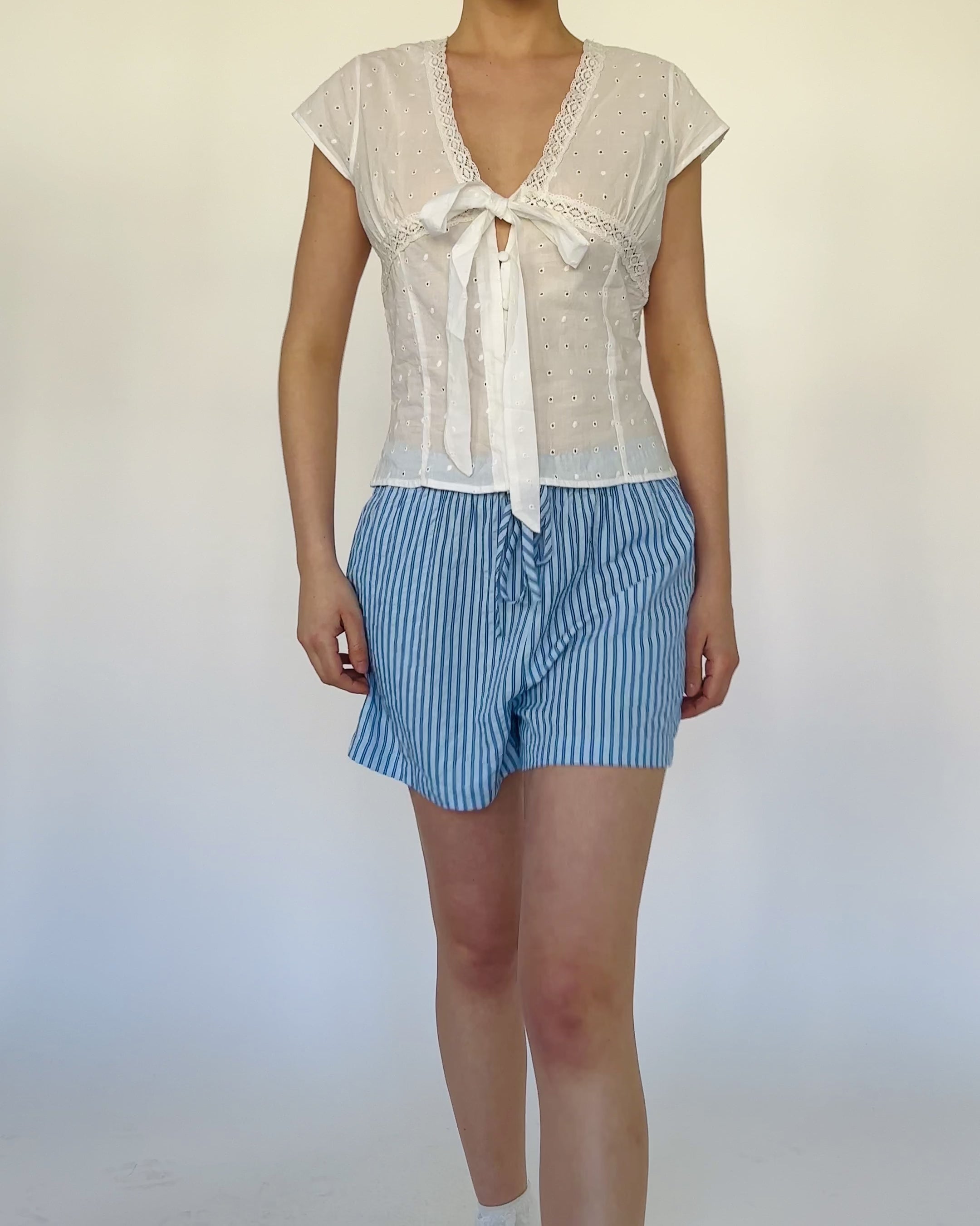 Model trägt eine luftige Shorts mit blauen Streifen und mit Bindebändern, elastischem Bund und weitem Schnitt zu einer weißen ärmellosen Bluse.