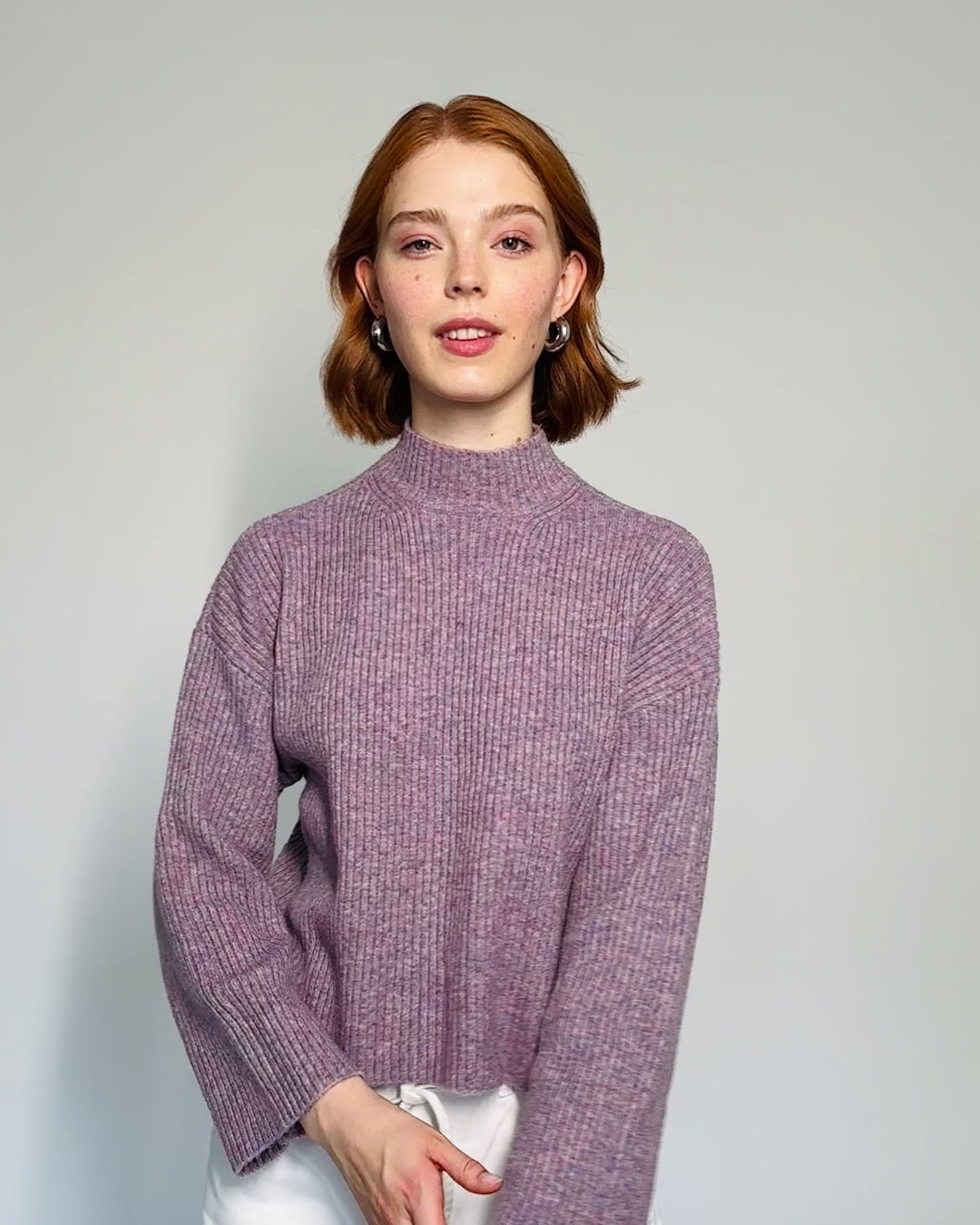 Model trägt einen gerippten lila Pullover mit Stehkragen zu einer weißen Jeans.